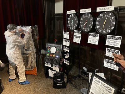 The setup of the exhibit: Mylar-covered chamber (with staffer in clean suit); the control unit; and the clocks held up on a shelf. Everything is covered in warning labels.