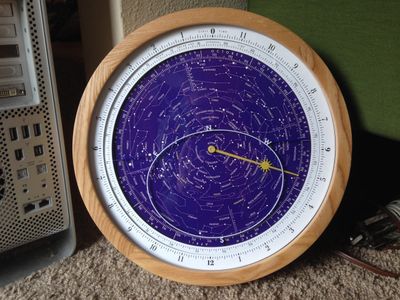 The completed clock (12-hour version). Wooden frame, white hour ring, and bluish-purple sky dial with stars inside, and a yellow hand pointing to the hour on the white ring and the date on the edge of the blue one.